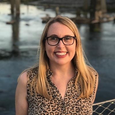 A white woman with blond hair and glasses, smiling toward the camera in front of a river